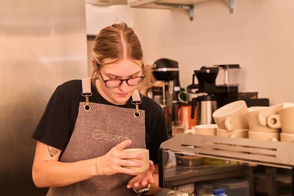 At Café Essenz, a woman in an apron pours coffee into a cup, exemplifying her expertise as a barista in a cozy setting.