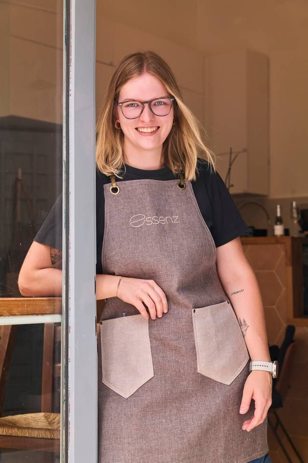 A woman wearing glasses and an apron stands by a window, representing her work as a barista at Café Essenz.