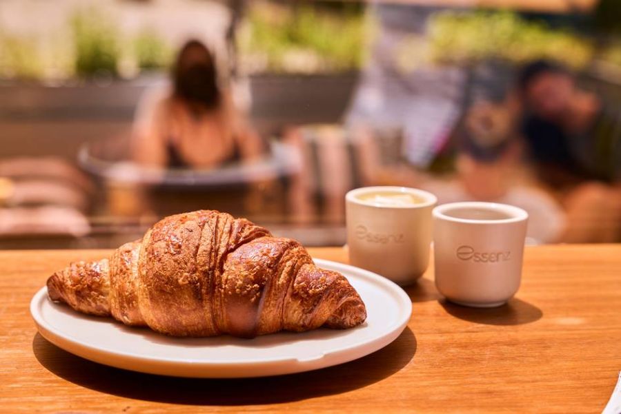 A golden-brown Essenz croissant with flaky layers, resting on a white plate, ready to be enjoyed with coffee.