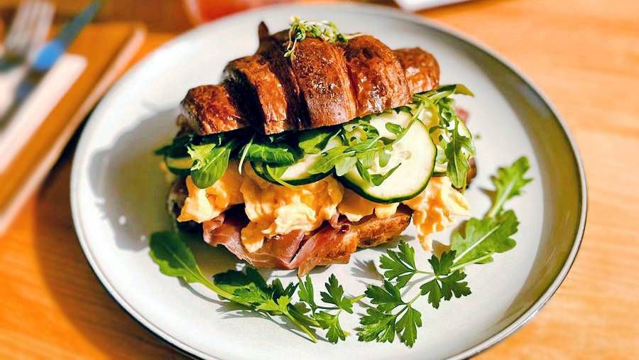 A white plate featuring a croissant stuffed with scrambled eggs vegetables, and cucumber.